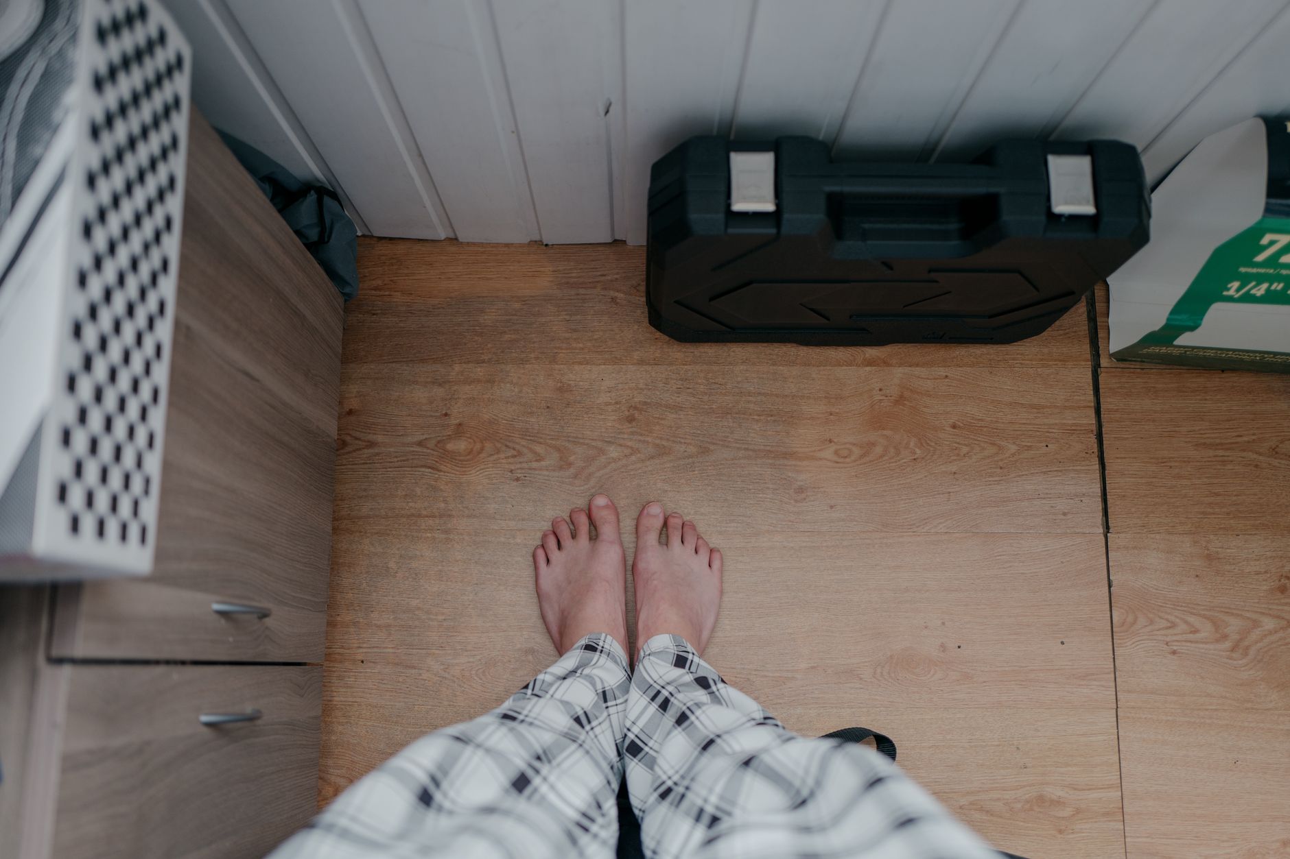 Draufsicht auf nackte Füße in Pyjamas auf einem Holzboden in einem Innenraum, daneben ein Werkzeugkasten.